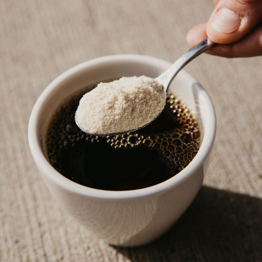 Hand holding a tablespoon of protein powder over a cup of black coffee.