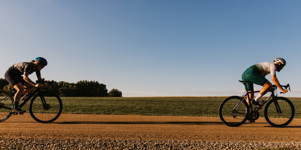 A male and female cyclist on a gravel road with green field and blue sky in the background.